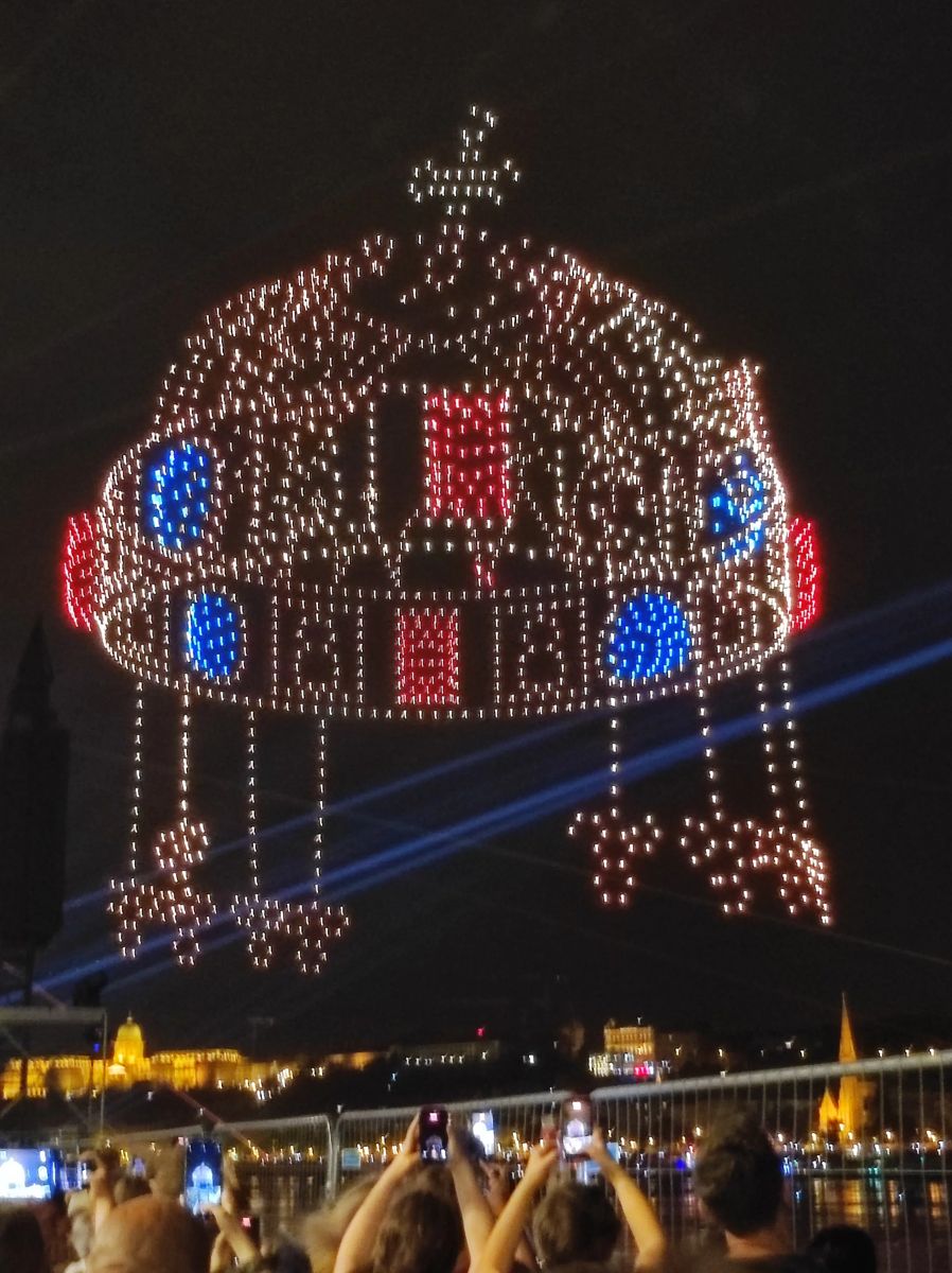 The Holy Crown of Hungary drawn in lights by drones above the Danube on Saint Stephen's Day, with the crowd watching from the riverbank