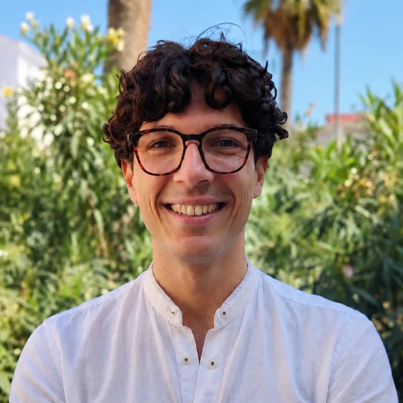 Portrait of Eros Reale smiling, in a white shirt, in a sunny garden, framed by the eight-petal ER flower